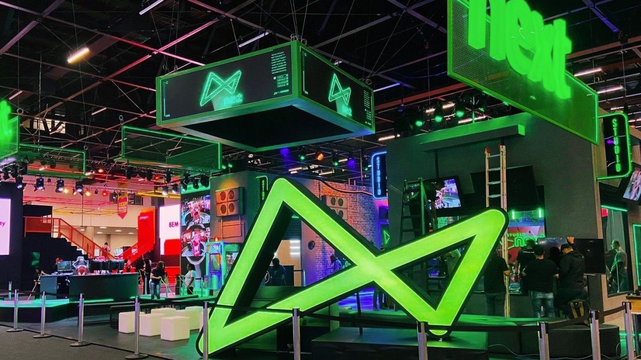 A imagem mostra o estande do next na CCXP num cenário majoritariamente verde, com o nome e diversos logos do next.