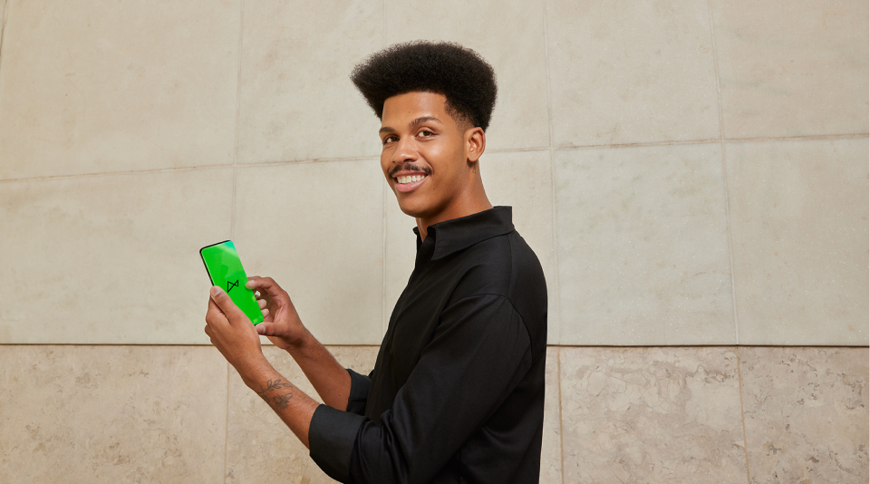 A imagem mostra um homem negro, de cabelo crespo e bigode, olhando pra câmera e vestindo uma camisa na cor preta. Ele está segurando o celular, mostrando sua tela com o logo do next.